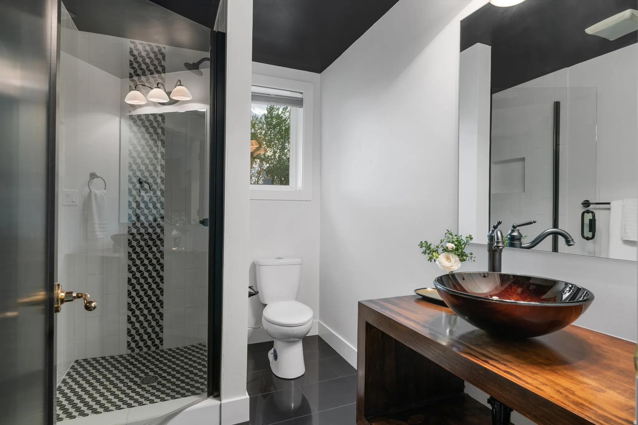 Contemporary bathroom featuring a glass-enclosed shower with chevron tile, a floating wood vanity with a vessel sink, dark flooring, and a window providing natural light