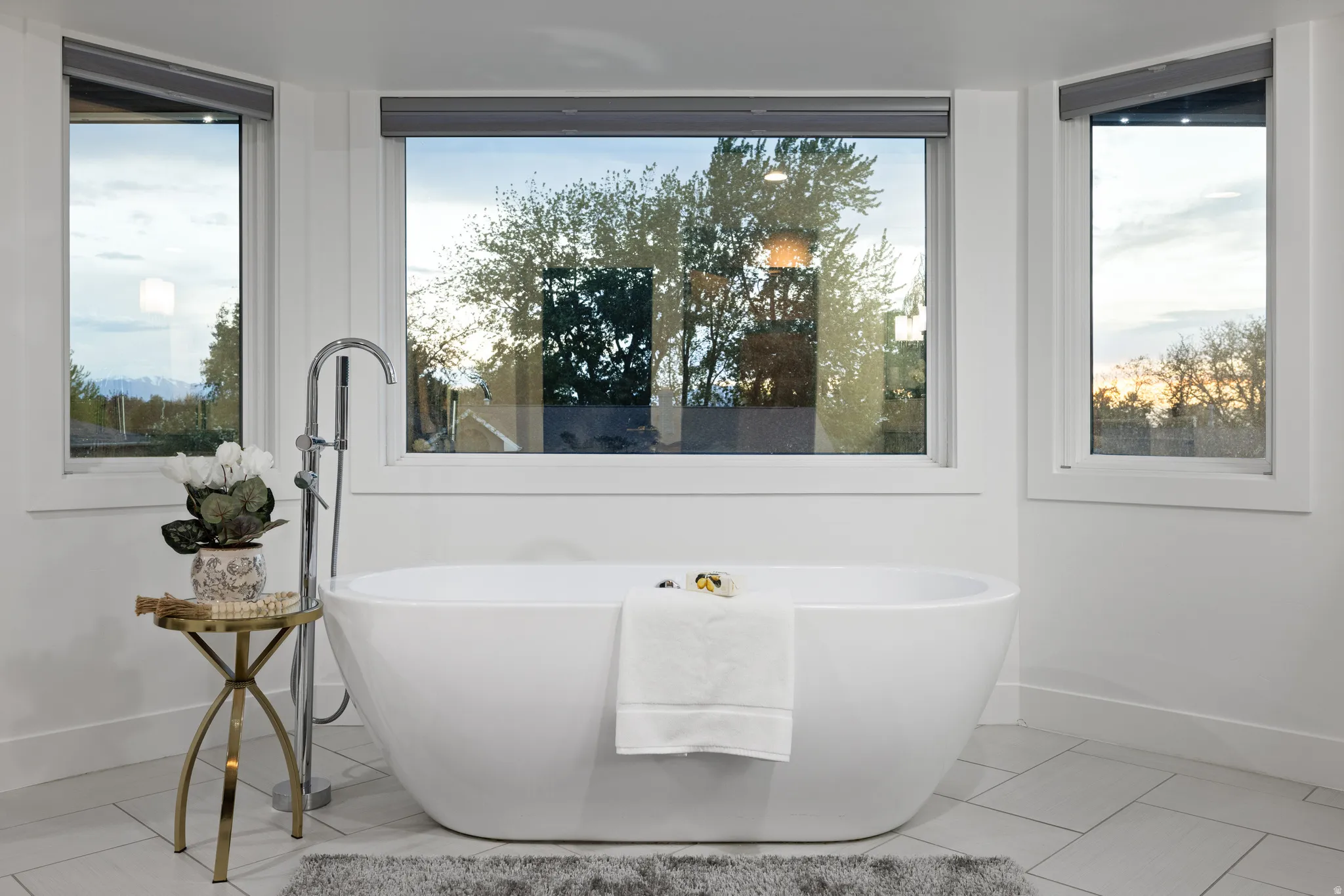 Freestanding soaking tub with chrome floor-mounted faucet, surrounded by large windows with roller shades