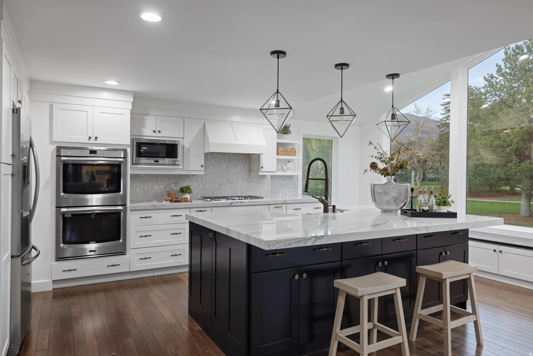 Kitchen featuring a dark-toned island with a light-toned countertop, white cabinetry, stainless steel appliances, wood-finish flooring, and a large window overlooking trees and a mountain