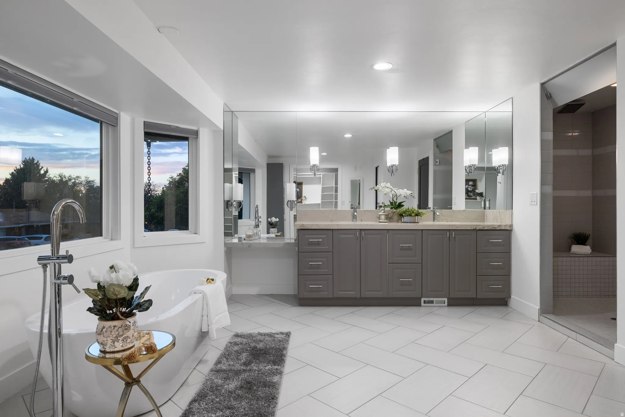 Freestanding soaking tub with floor-mounted faucet, dual vanity with undermount sinks and extensive countertop space, integrated shower with tiled bench seating, multiple windows, and large format tile flooring