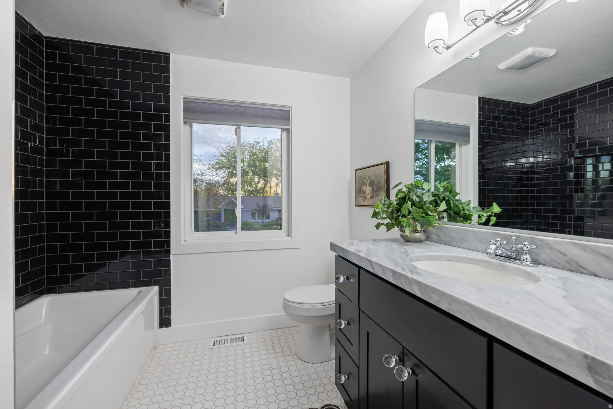Bathroom featuring a black subway tile shower surround, white ceramic bathtub, white hex tile flooring, white window, and a dark wood vanity with a light stone countertop and a chrome faucet