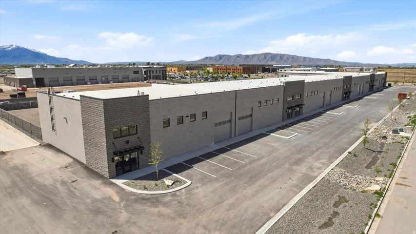 Modern commercial building featuring a two-tone facade with a prominent brick section, multiple roll-up doors, an expansive paved parking lot, and a manicured landscape bed at the main entrance