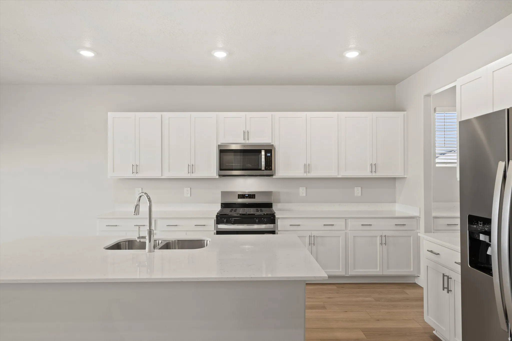 Kitchen featuring an island with an undermount sink, white shaker-style cabinetry, stainless steel appliances, light-toned wood-finish flooring, and recessed ceiling lighting