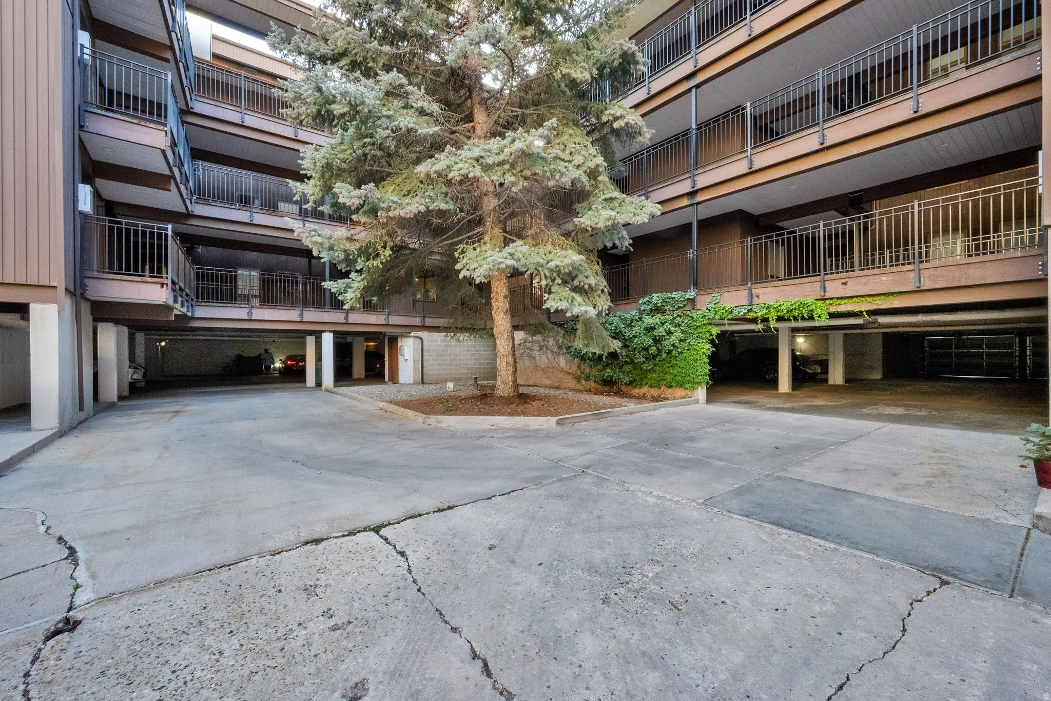 Multi-level apartment building featuring private balconies with black metal railings, a central parking area, mature landscaping, and an integrated covered parking structure