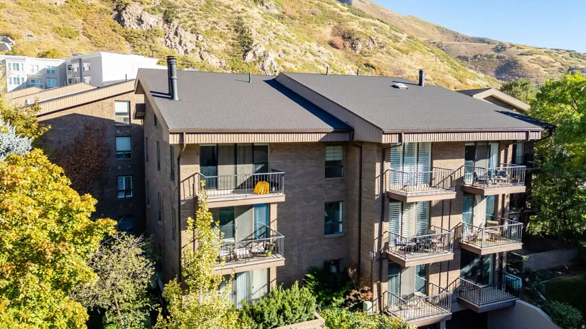 Multi-story building featuring brick and vertical siding facades, dark asphalt shingle roofing, multiple private balconies with metal railings, and a mountainous backdrop