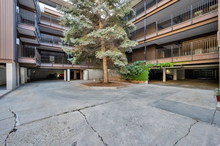 Multi-level apartment building featuring private balconies with black metal railings, a central parking area, mature landscaping, and an integrated covered parking structure