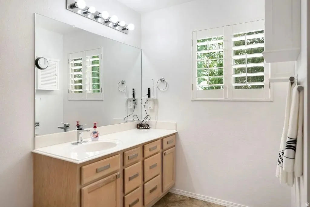 Bathroom featuring a light wood-finish vanity with dual sinks, a large wall mirror, and a window with white plantation shutters