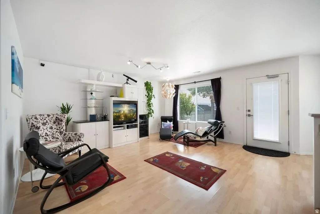 Spacious living area featuring light wood-finish flooring, a built-in media console with storage, a large window, a track lighting fixture, and a white door with an integrated blind