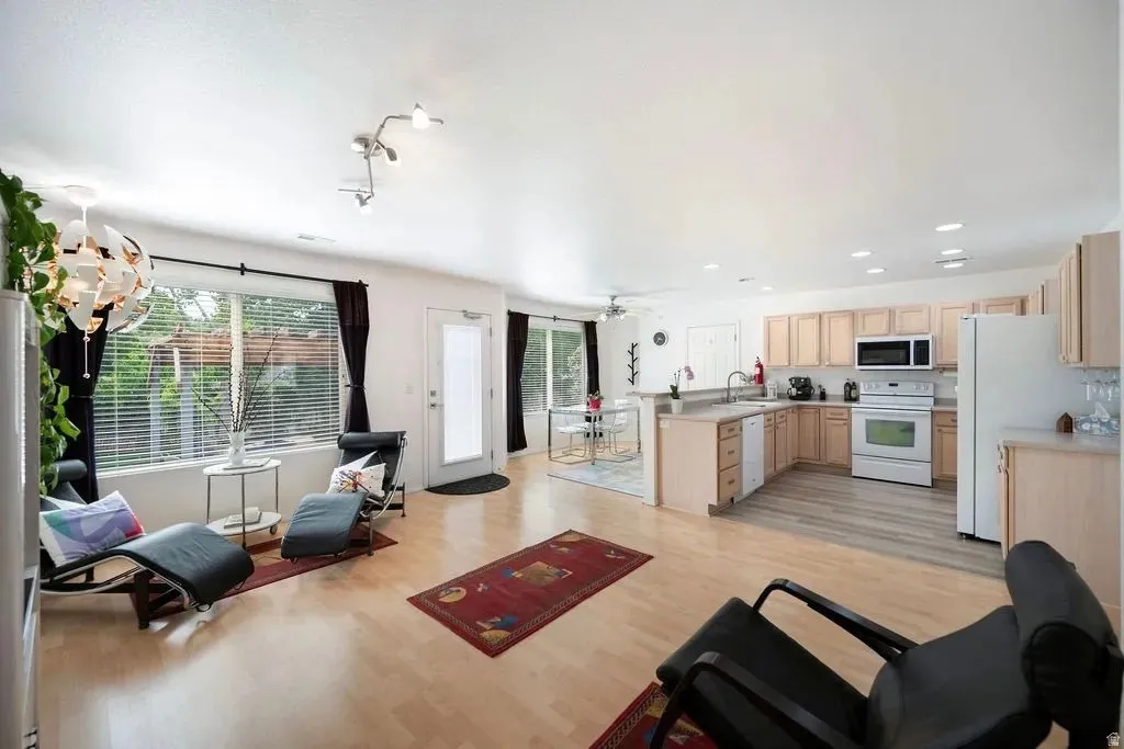 Open-concept living area with light wood-finish flooring, a contemporary overhead light fixture, and a kitchen featuring light wood cabinetry, white appliances, and a breakfast bar