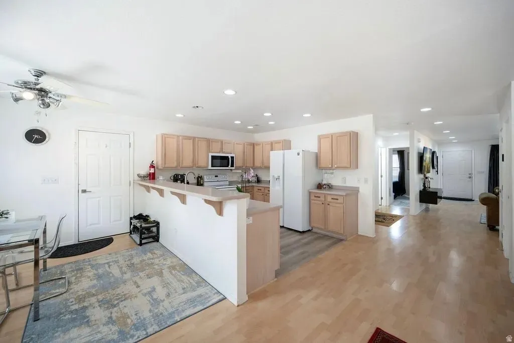 Open-concept kitchen featuring light wood cabinetry, white appliances, recessed lighting, and a breakfast bar