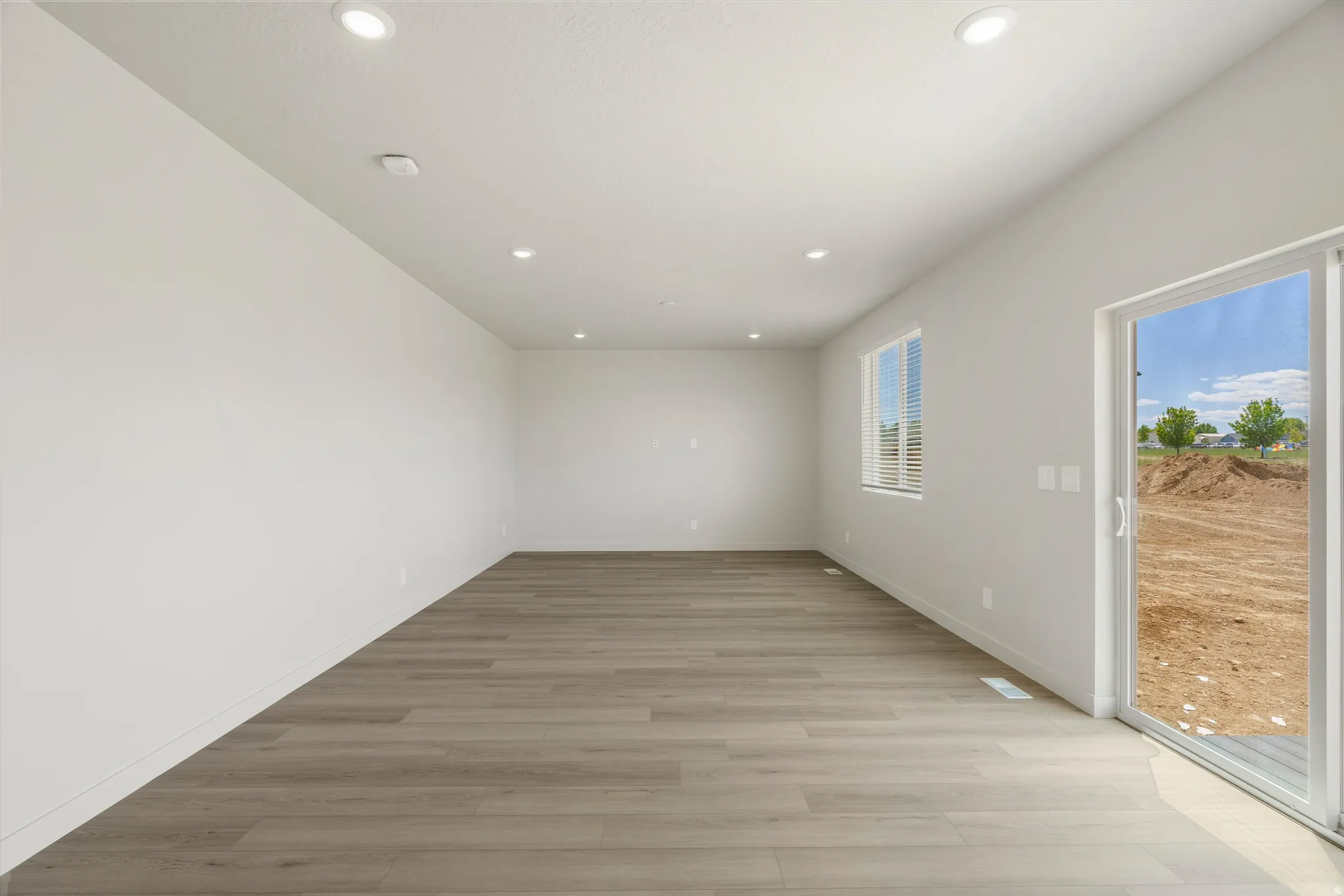 Spacious room featuring light wood-finish flooring, recessed lighting, a slider door, and a horizontal window with blinds