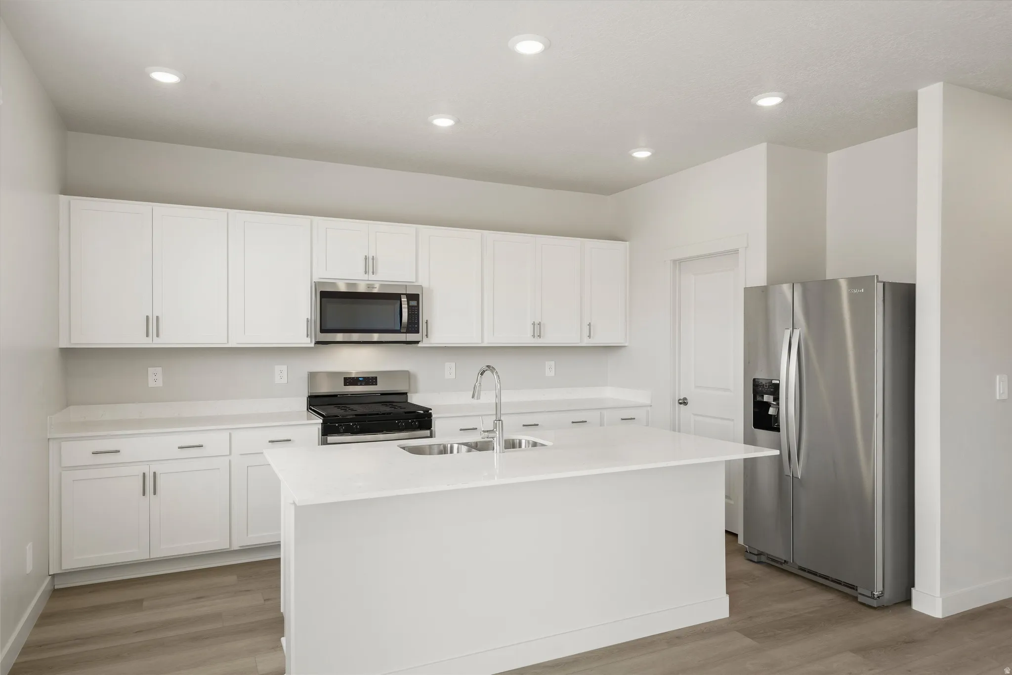 Contemporary kitchen featuring an island with an undermount sink, white cabinetry, integrated stainless steel appliances, light-toned countertops, and wood-finish flooring