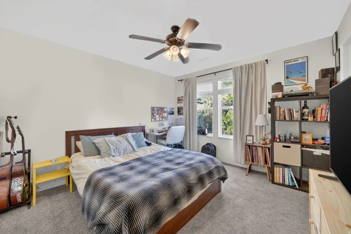 Carpeted bedroom with a ceiling fan light fixture, white walls, and a large window with white blinds and light-colored curtains