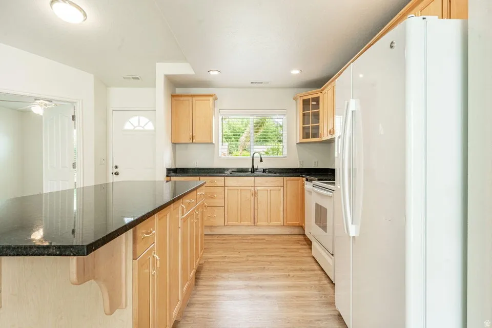 Kitchen featuring a large island with dark stone countertops, light wood cabinetry, wood-finish flooring, white appliances, and a window above the sink
