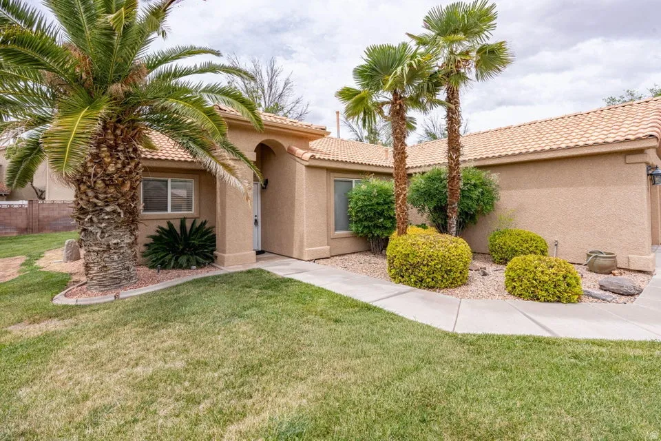 Exterior stucco residence featuring a concrete tile roof, mature palm trees, desert landscaping with gravel beds, established shrubs, and a concrete walkway