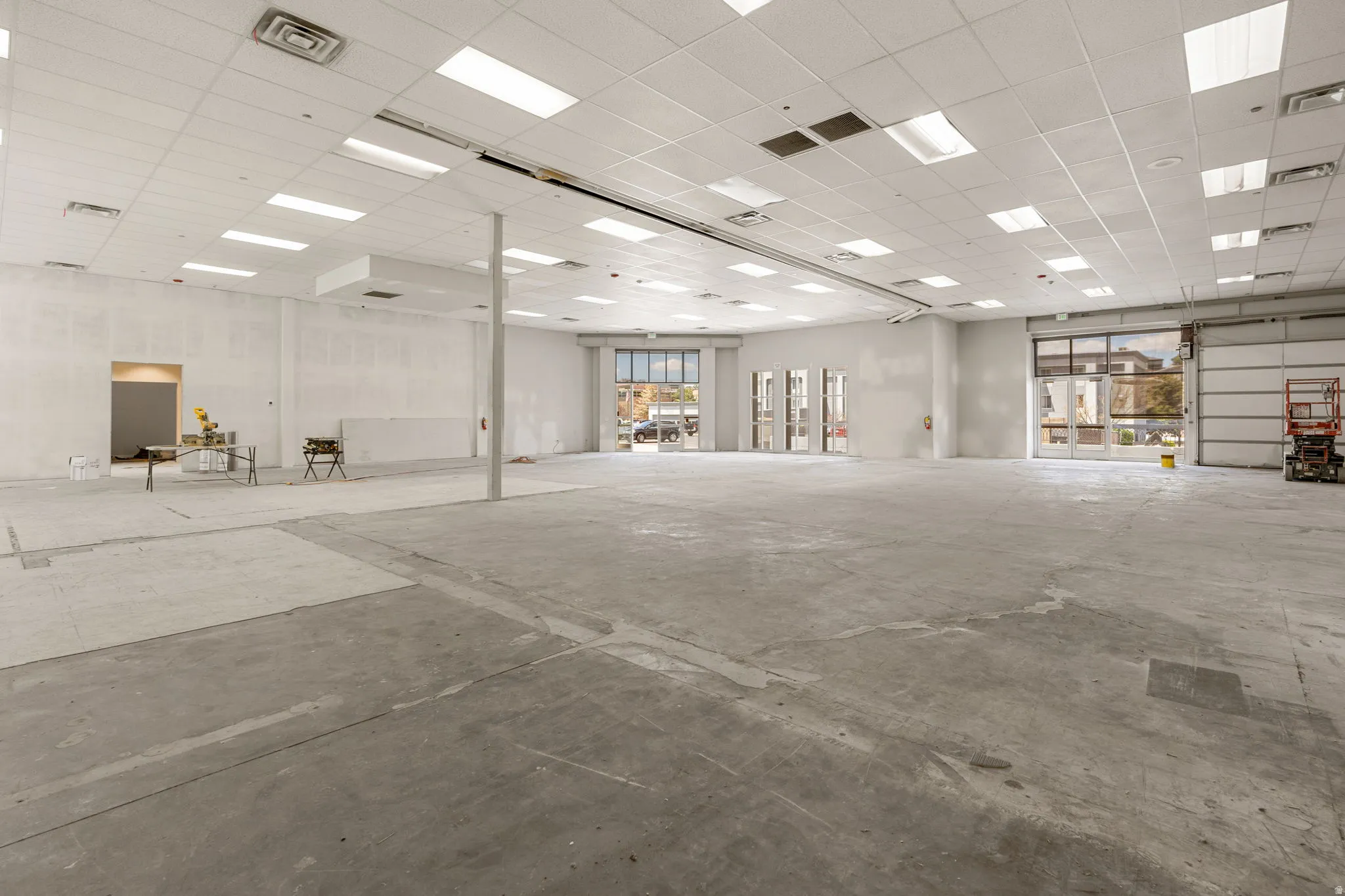 Expansive commercial space featuring a concrete floor, drop ceiling with integrated lighting, multiple storefront windows, and a large roll-up door