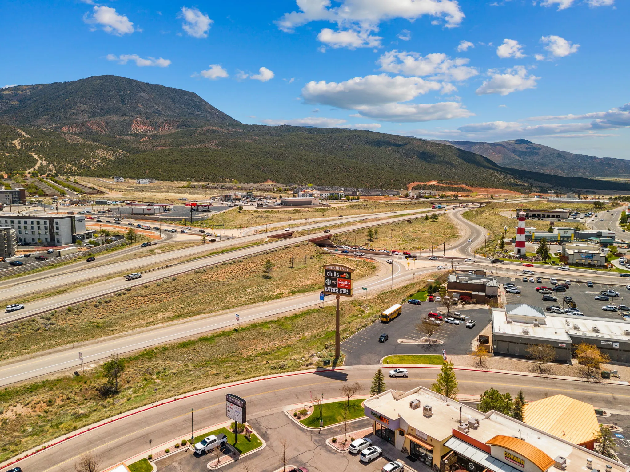 Expansive commercial area featuring multiple retail buildings, a prominent highway interchange, parking facilities, and surrounding natural mountainous terrain
