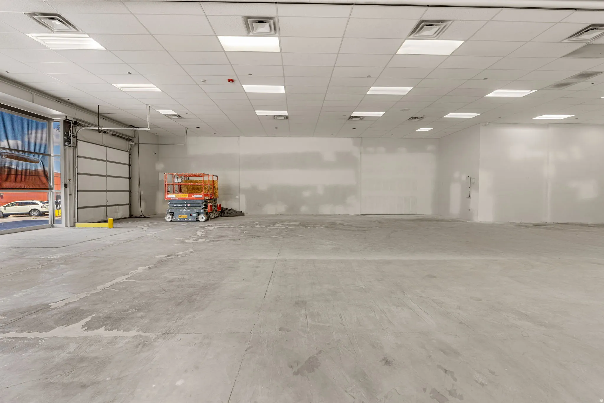 Spacious commercial interior featuring a roll-up garage door, concrete flooring, and a grid ceiling with integrated lighting