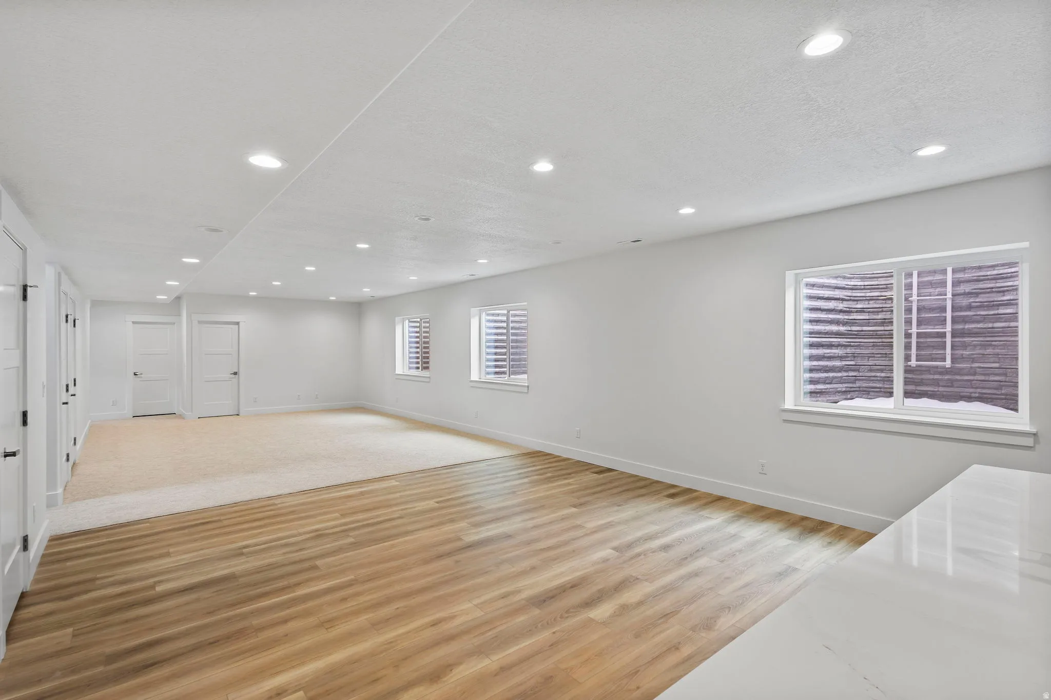 Spacious room featuring wood-finish flooring and neutral carpet, white walls, multiple recessed lights, and several white doors with black hardware
