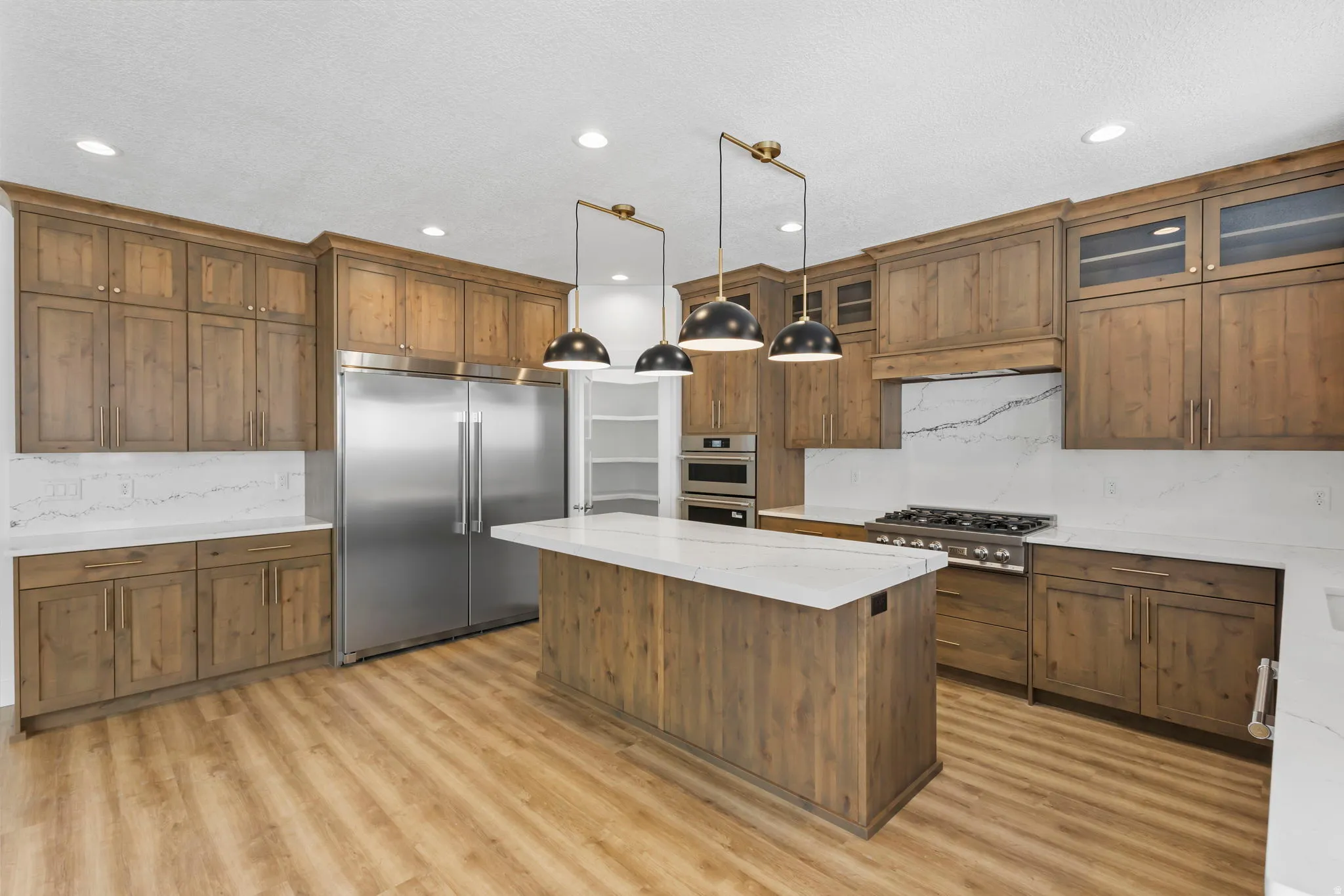 Open-concept kitchen featuring a central island with a white countertop, wood-finish flooring, full-height cabinetry with crown molding, stainless steel appliances, and ceiling-mounted pendant lighting