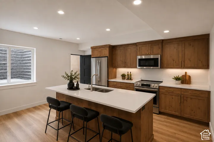 Well-appointed kitchen featuring light wood-finish flooring, a central island with a double basin sink, white countertops, stainless steel appliances, and wood cabinetry with under-cabinet lighting