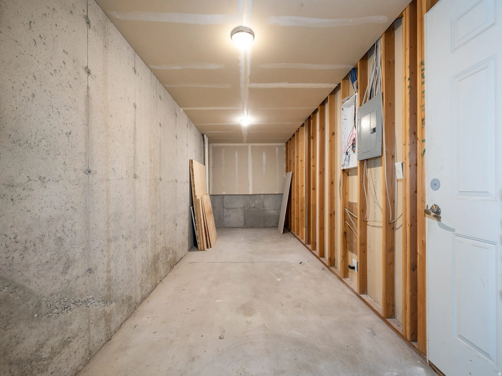 Unfinished basement space featuring a concrete floor and walls, exposed wood framing, an electrical panel, and a white door with hardware