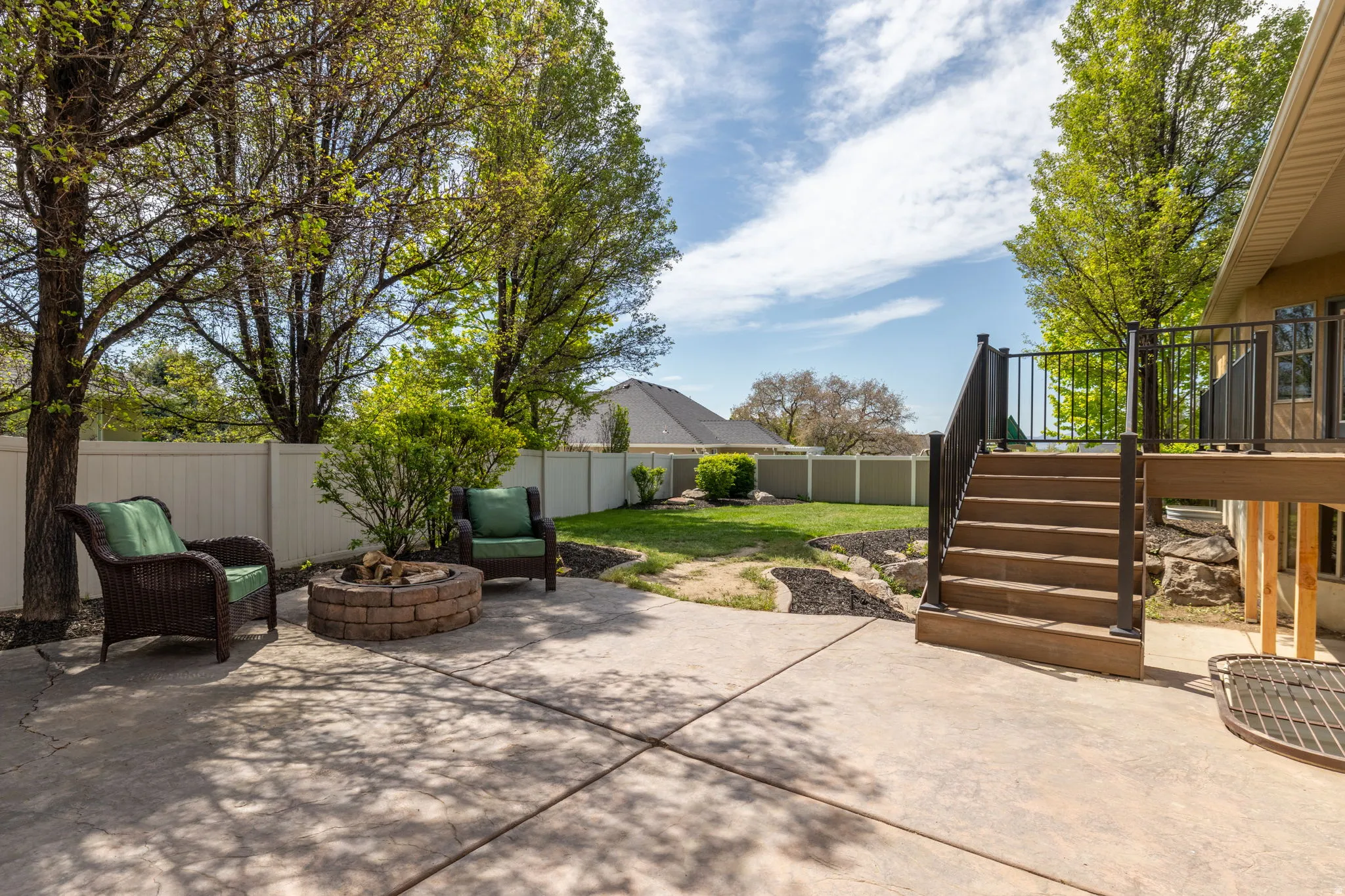 Expansive concrete patio featuring a built-in stone fire pit, a multi-level composite deck with dark railings, a manicured lawn, and mature trees providing shade