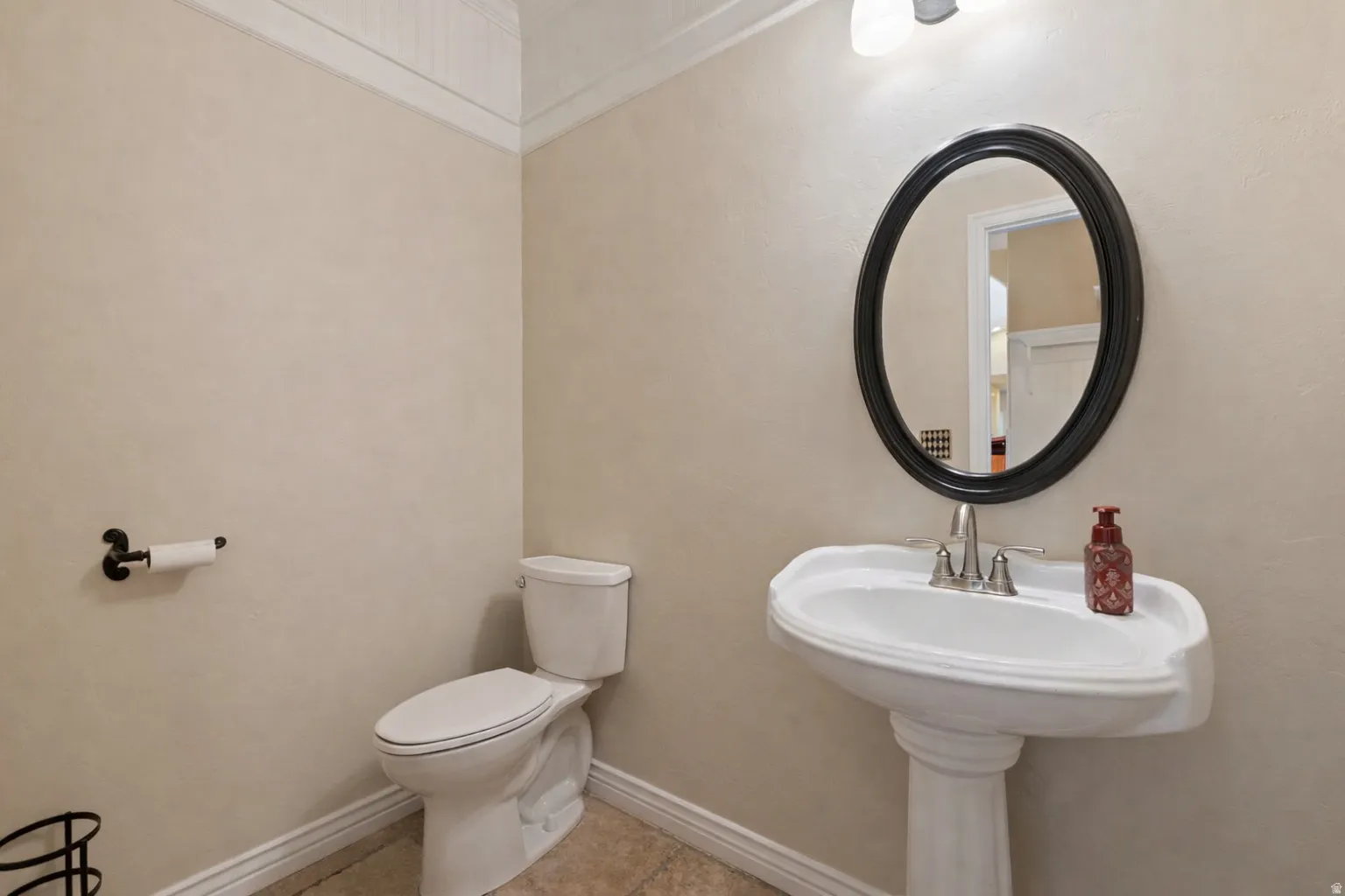 Pedestal sink with an oval-framed mirror, complemented by a two-piece toilet, tiled flooring, and light-toned walls