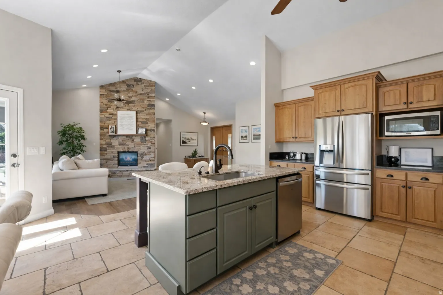 Open-concept kitchen featuring light-toned tile flooring, a central island with a sink and light-colored countertop, wood cabinetry, and stainless steel appliances