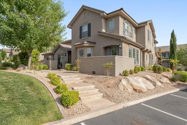 Stucco exterior featuring multiple windows with dark shutters, a front entry with concrete steps, and integrated landscaping with mature trees and decorative rocks