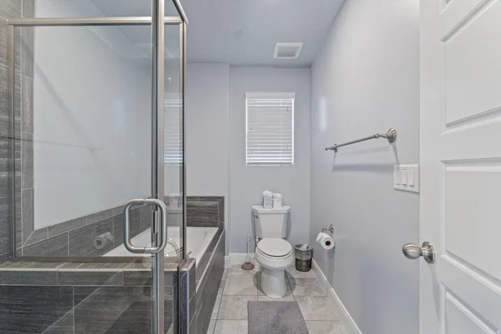 Full bathroom featuring a glass-enclosed shower-tub combination with dark gray tile surround, light gray wall paint, a white toilet, and window with blinds