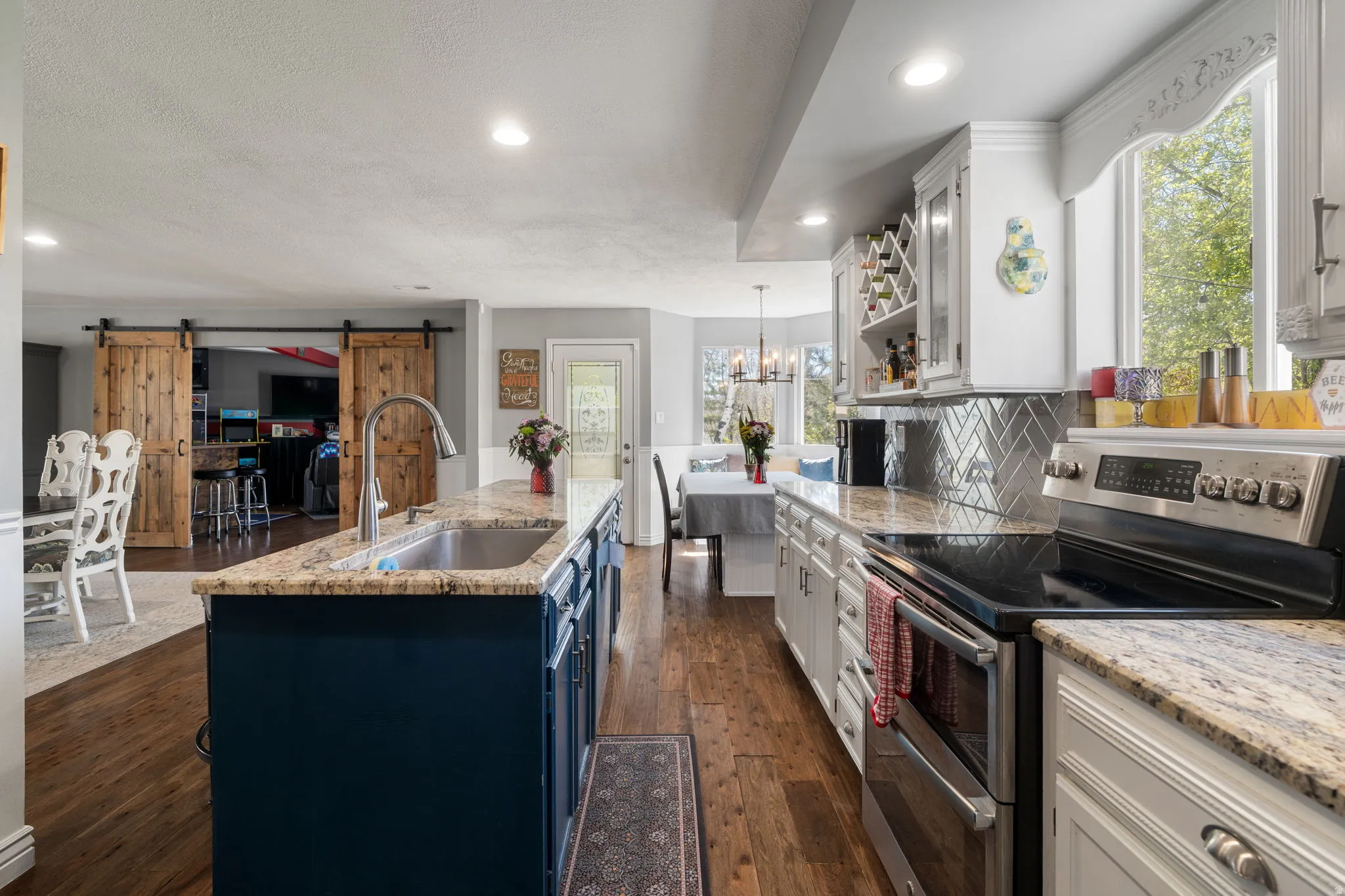 Gourmet kitchen featuring wood-finish flooring, a central island with a stainless steel sink and granite countertop, white cabinetry, a herringbone tile backsplash, and stainless steel appliances