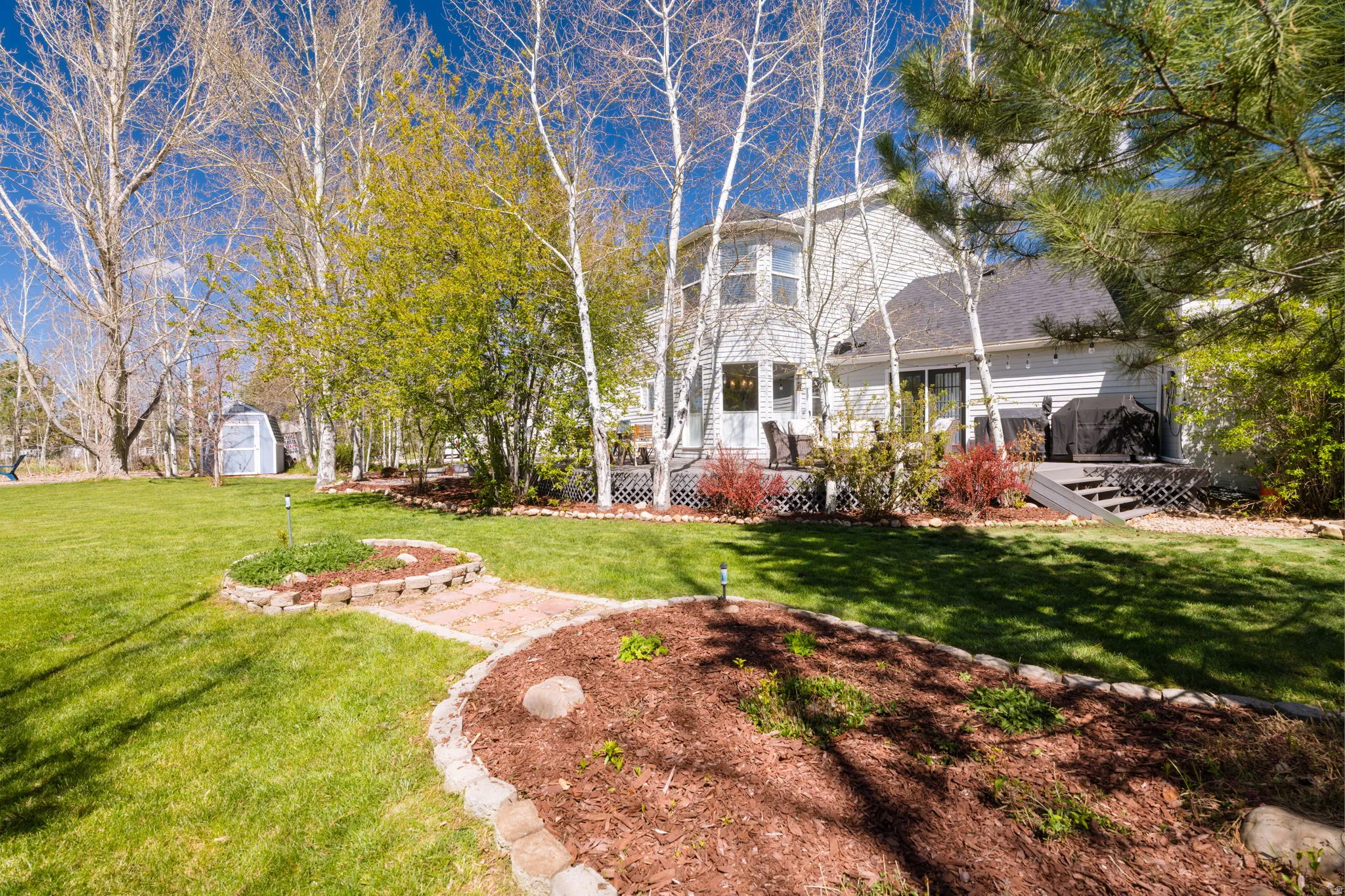 Expansive backyard featuring a tiered deck, mature trees, landscaped garden beds with stone edging, a stone-paved pathway, and a detached shed