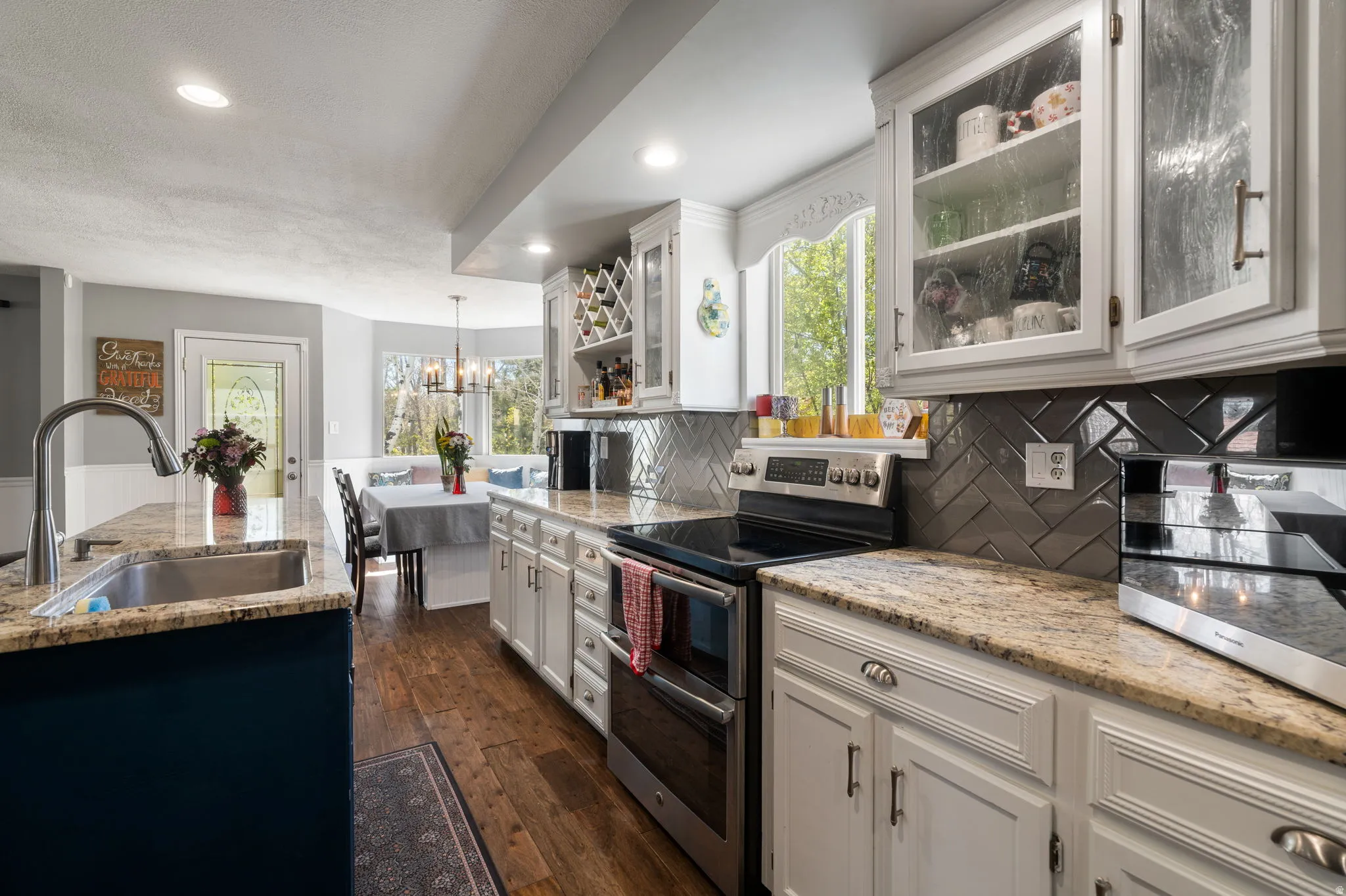 Kitchen featuring a stainless steel sink, granite countertops, white cabinetry with glass-front uppers, a herringbone tile backsplash, and wood-finish flooring