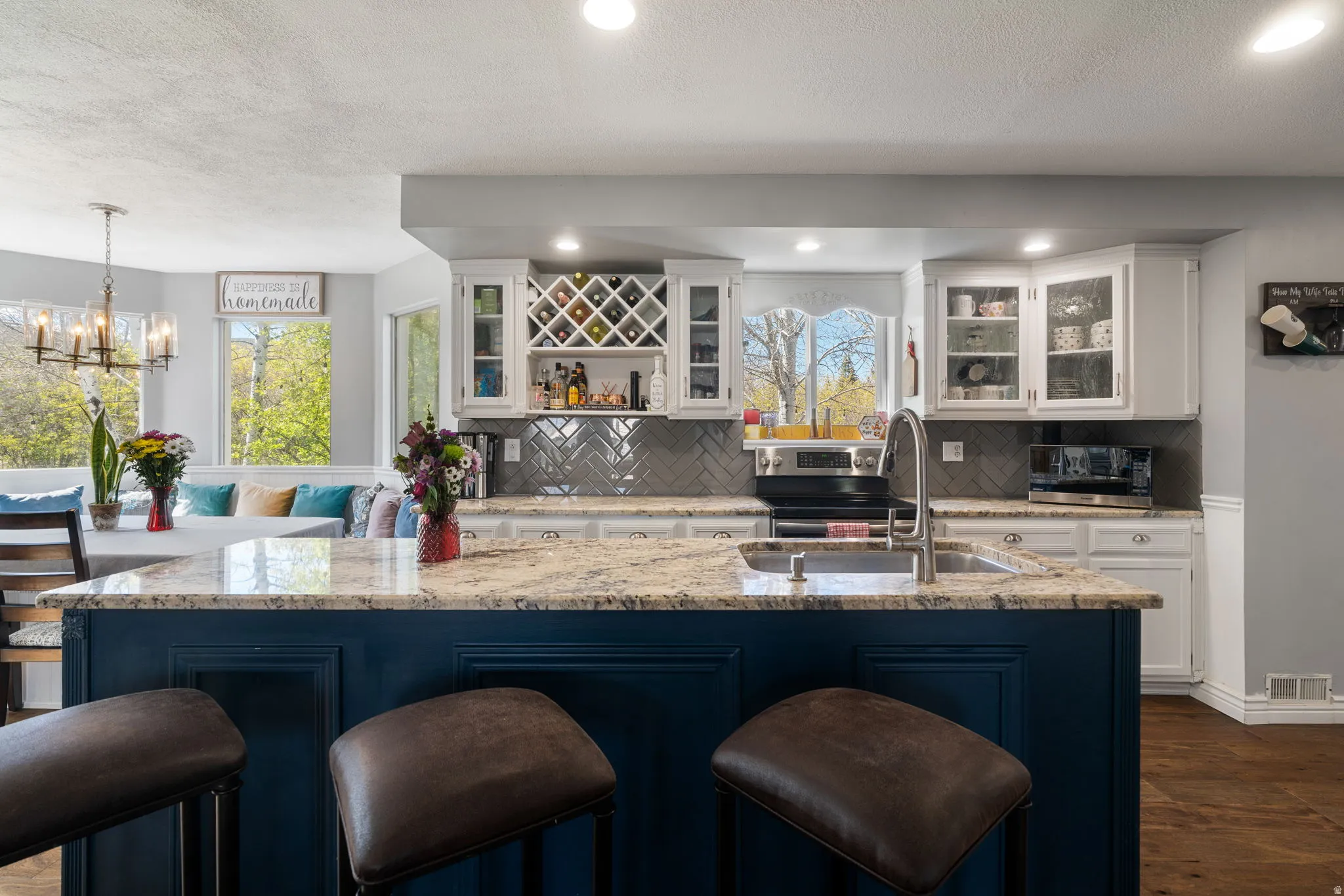 Kitchen featuring a blue-painted island with a built-in sink and stainless steel faucet, granite countertops, white cabinetry with glass-front accents, and a grey herringbone tile backsplash