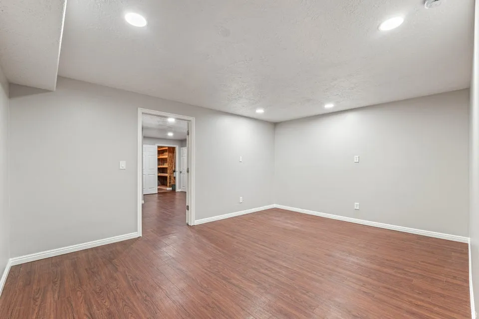 Spacious room featuring wood-finish flooring, light gray walls, white baseboards, recessed lighting, and a doorway to an additional storage area with built-in shelving
