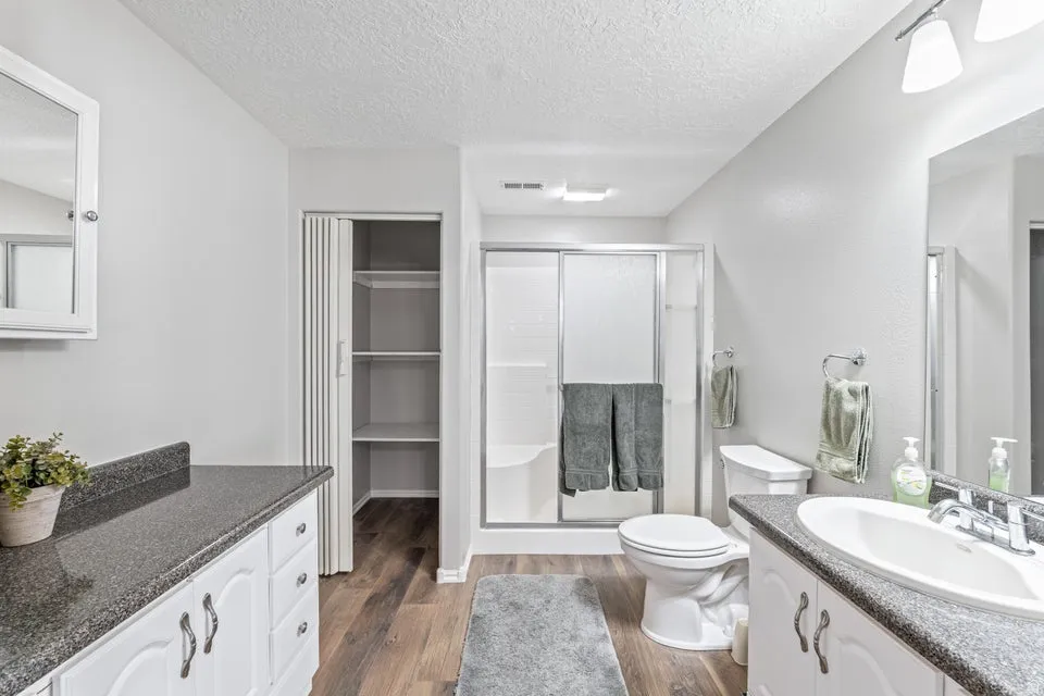 Bathroom featuring wood-finish flooring, a white vanity with a dark speckled countertop, a toilet, a shower with sliding glass doors, and an integrated storage closet with shelving