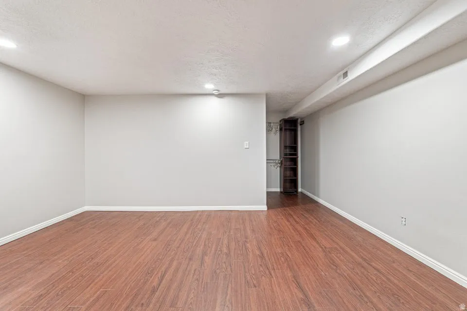 Spacious room featuring wood-finish flooring, recessed ceiling lighting, light gray wall paint, white baseboards, and built-in shelving