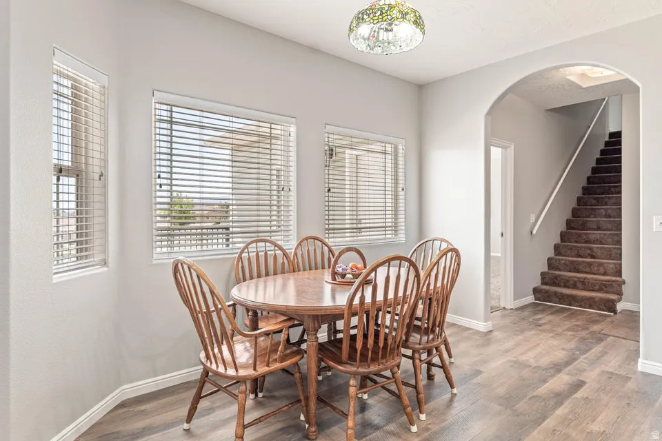 Dining area featuring wood-finish flooring, multiple windows with horizontal blinds, and an arched doorway leading to a carpeted staircase