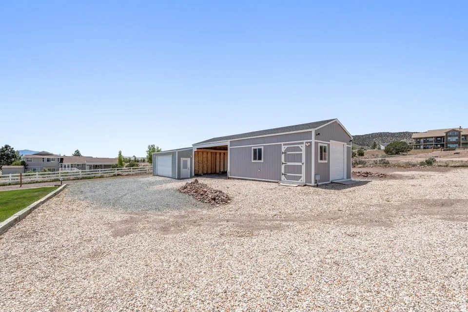 Gravel lot featuring a detached garage with a white overhead door, an attached open-sided carport, a separate storage shed, and a white picket fence bordering a grassy area