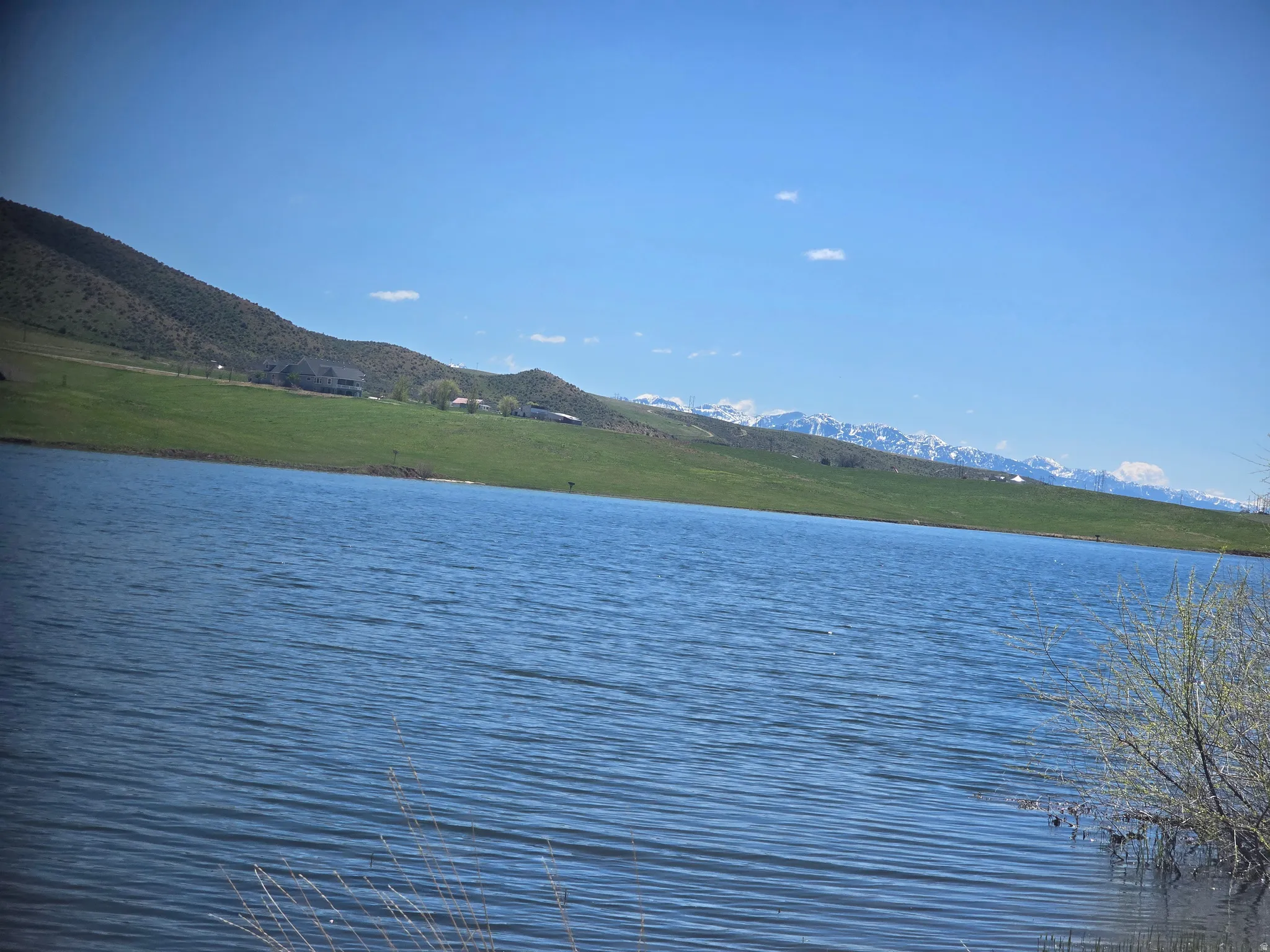 Tranquil waterfront setting featuring a body of water, green hillsides, distant snow-capped mountains, and a clear blue sky