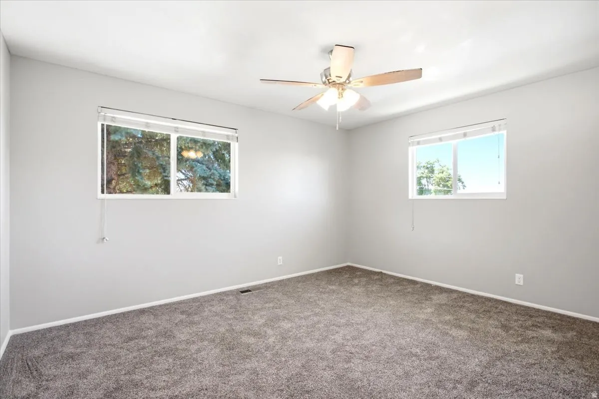 Spacious room featuring neutral carpeting, two large windows with blinds, a ceiling fan with integrated lighting, and light gray wall tones
