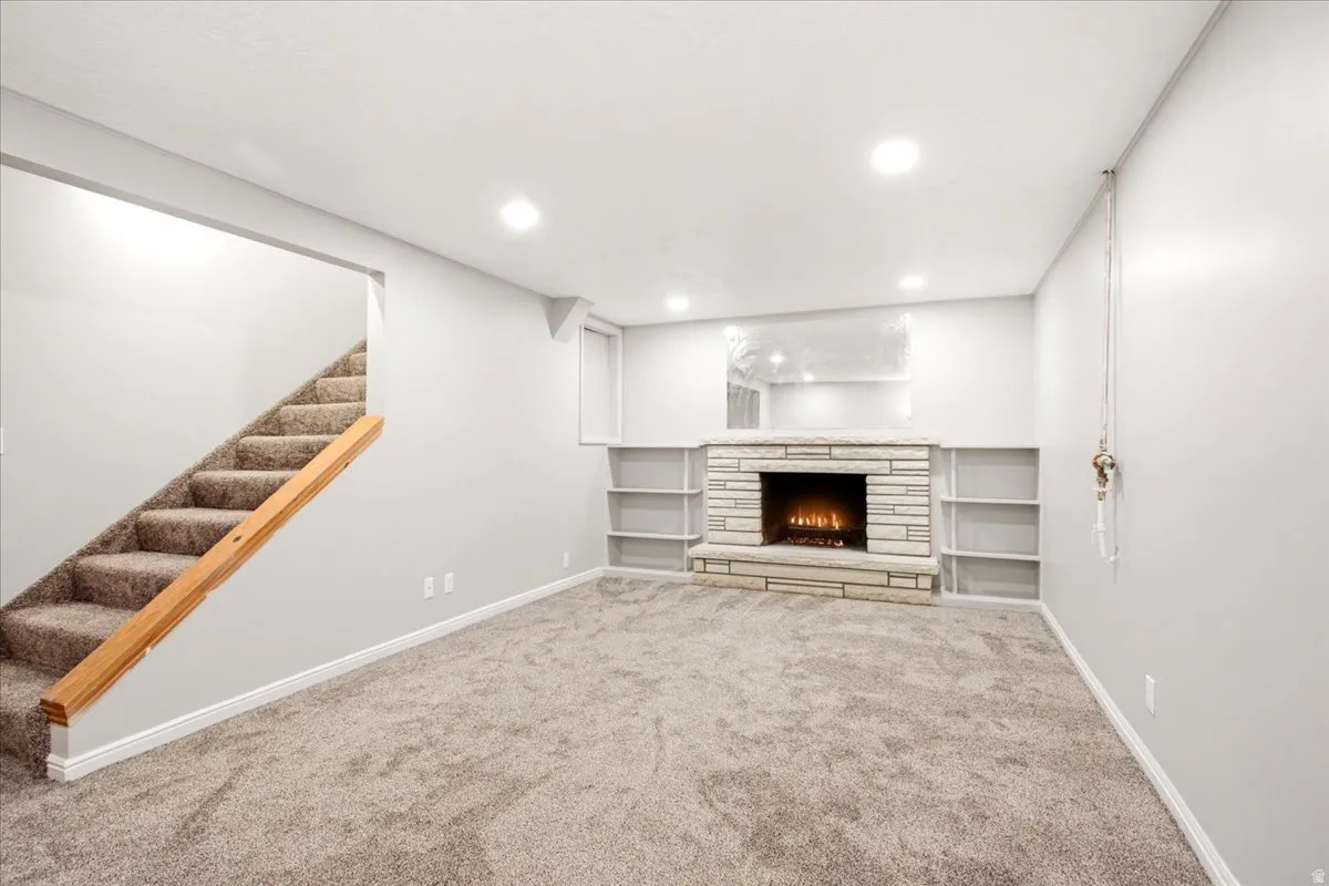 Carpeted room featuring a stone fireplace with a hearth, built-in shelving, recessed lighting, and a carpeted staircase with a wood handrail