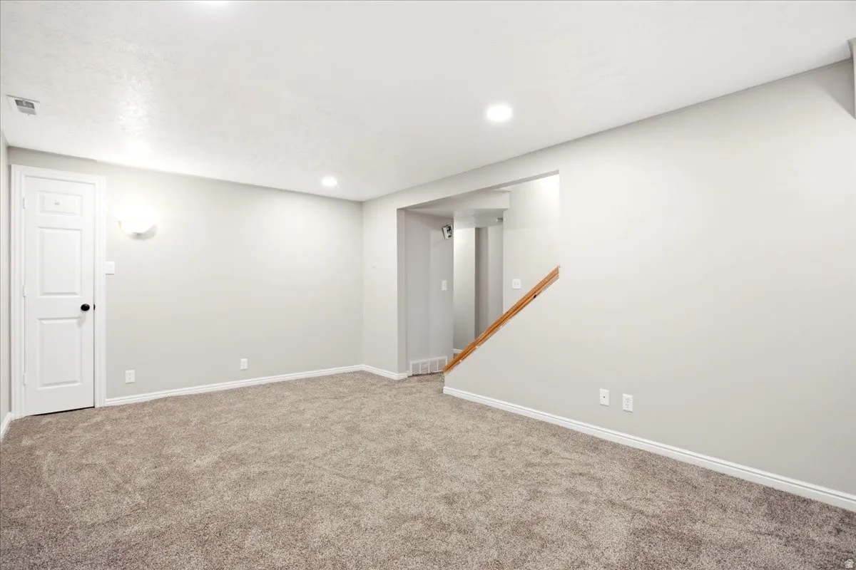 Spacious room featuring neutral carpet flooring, recessed lighting, a white panel door, and a wooden stair railing