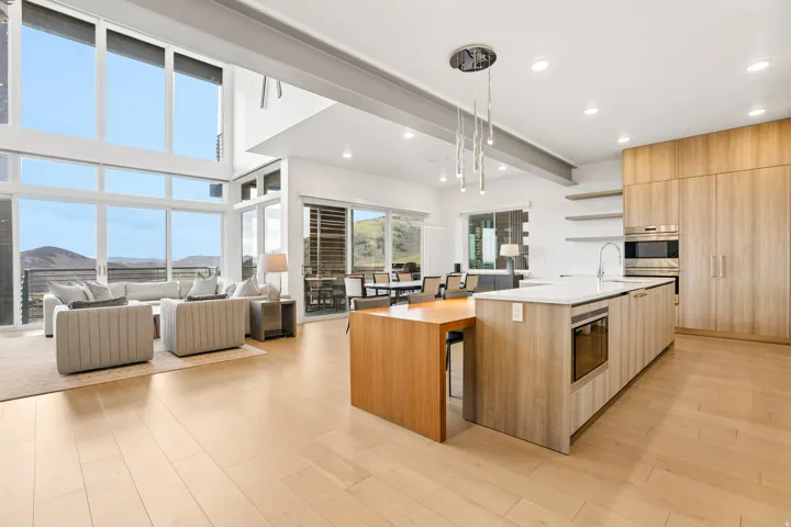 Modern open-concept living area featuring a kitchen island with integrated dining, light wood-finish flooring, and a two-story window wall