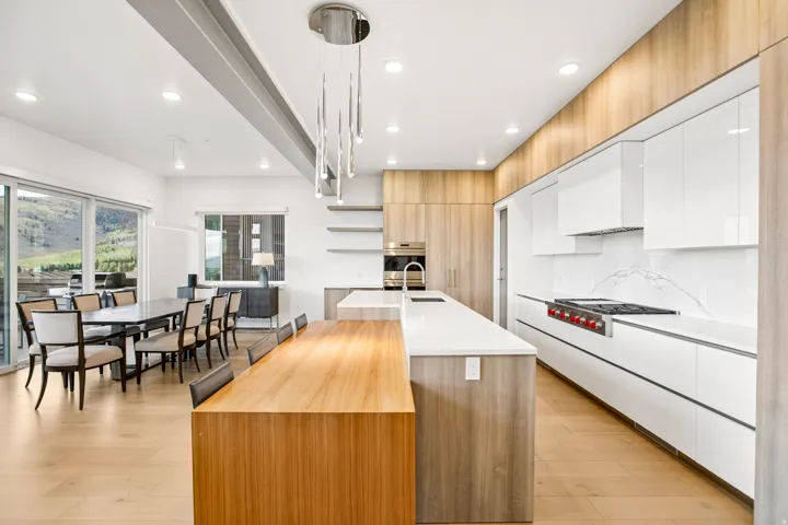 Gourmet kitchen and dining area featuring a lengthy island with wood-finish paneling, light wood-finish flooring, white flat-panel cabinetry, a stainless steel range, and a full-height marble backsplash
