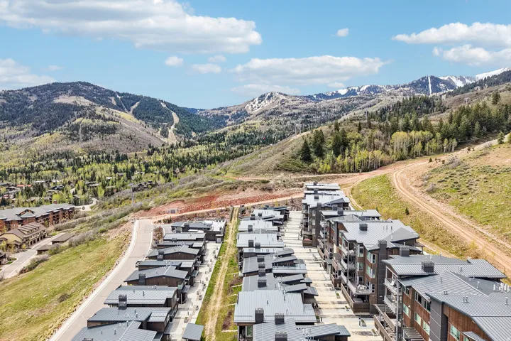 Contemporary multi-story buildings featuring dark paneling and balconies, situated amidst rolling hills with visible ski slopes and snow-capped peaks