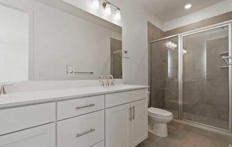Bathroom featuring a double vanity with white cabinetry and a light-toned countertop, a frameless glass shower enclosure with tiled walls, and recessed lighting