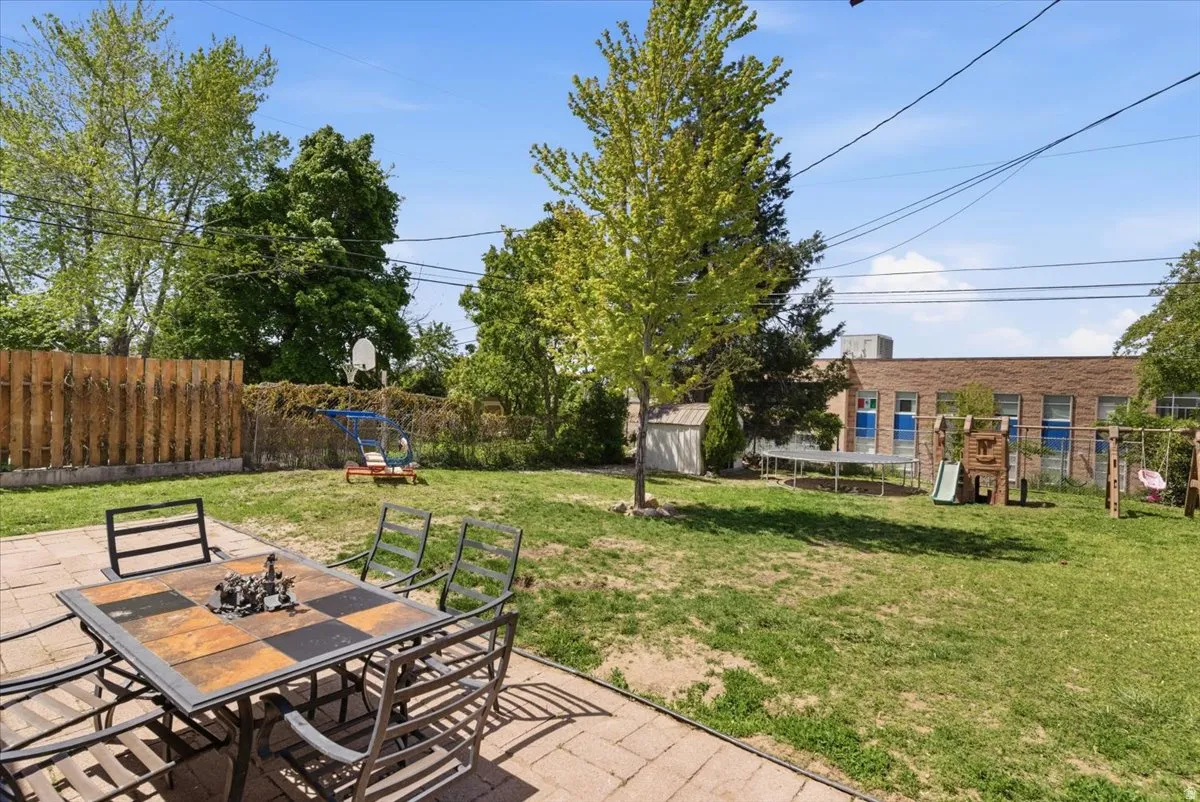 Expansive backyard featuring a paver patio with a checkerboard-patterned tabletop, a lush lawn, mature trees, a wooden privacy fence, and a white storage shed