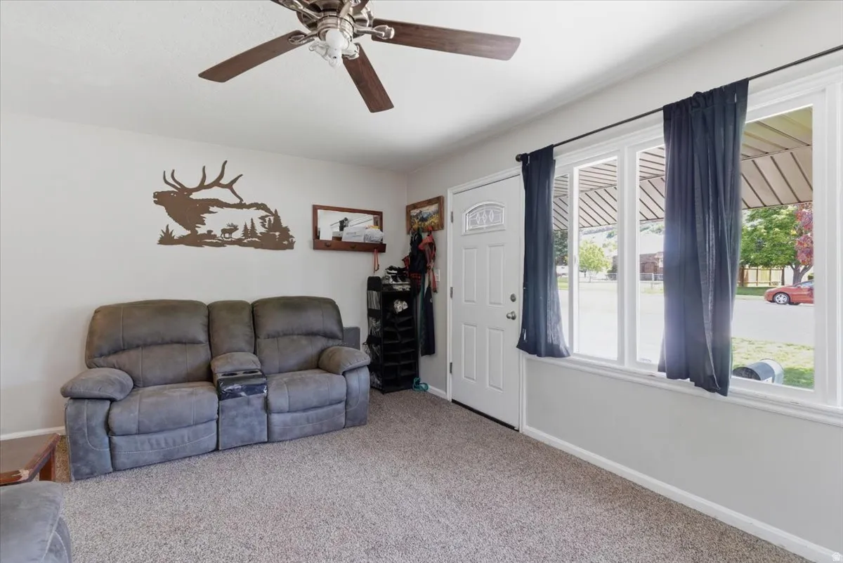 Living space featuring light-toned carpeting, a ceiling fan with wood-finish blades, and white walls